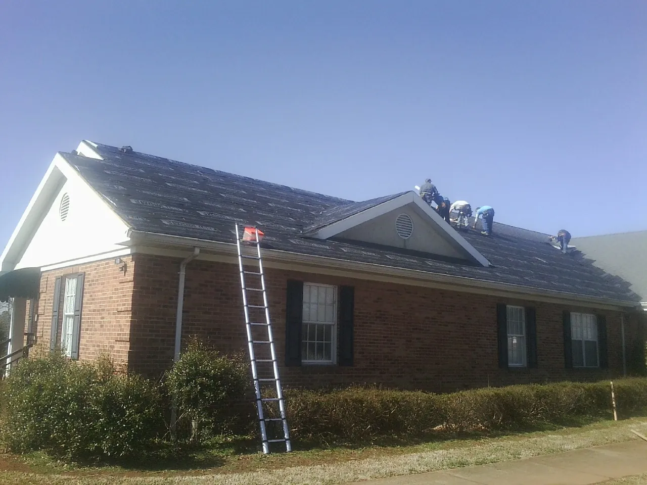 Crew tearing shingles off a brick ranch roof with a ladder set — Metal Roof Repair work by our crew in Opelika
