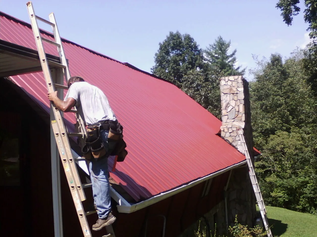 Roofer on a ladder beside a red metal roof and stone chimney — Soffit & Fascia Repair by Roofing Brothers in Opelika